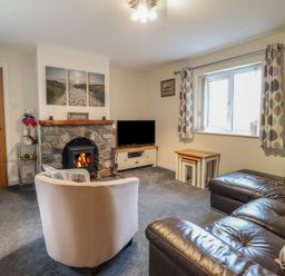 A living room with a stone fireplace a TV a brown leather sofa a white armchair and a window with curtains at Ysgubor Newydd Parc Isaf in Dyffryn Ardudwy