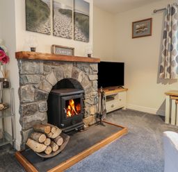 A living room with a stone fireplace and wood burning stove a TV on a stand and a small table with flowers at Ysgubor Newydd Parc Isaf in Dyffryn Ardudwy