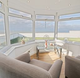 A sunroom with large windows overlooking a lawn and sea with two armchairs and a glass coffee table at The Gables in Tywyn