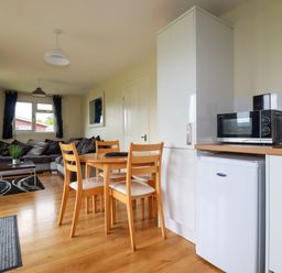A kitchen and living area with a wooden dining table and chairs a microwave on countertop a gray sofa and window with curtains at Gwanda in Tregolds near St Merryn