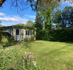 A garden with a green lawn flowers a tree and a small white building covered with plants at October House Holiday Cabin in Damerham