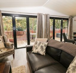 A living room with leather sofa patterned armchair glass dining table and large sliding doors overlooking a wooden deck at Somerford Lodge Langdale 7 White Cross Bay near Windermere