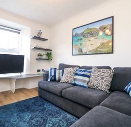 A sitting room with a dark grey sofa with patterned cushions a blue rug a television on a wall-mounted shelf and a framed coastal picture on the wall at Savoy View in Mullion
