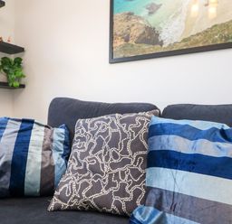 A black sofa with three patterned cushions and shelves with plants and jars on a white wall at Savoy View in Mullion