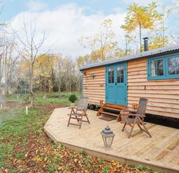 A wooden cabin with teal doors and windows on a wooden deck with two chairs and a lantern surrounded by trees and grass at Catchwater in Keal Cotes near East Kirkby