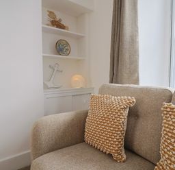 A beige sofa with textured cushions next to a built-in shelf with nautical decorations and a window with curtains at 47 Church Street in Lyme Regis
