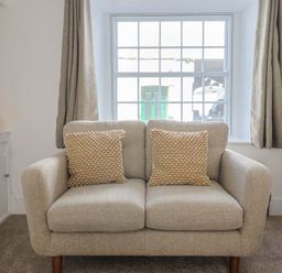 A beige loveseat with two textured cushions in front of a window with curtains and a lamp on the side at 47 Church Street in Lyme Regis