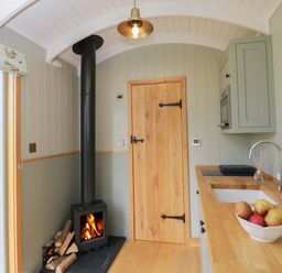 A kitchen with a wood burning stove next to a wooden door and a countertop with a sink and bowl of apples at Goose Whistle Hut in Foxham