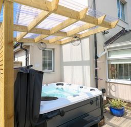 An outdoor hot tub under a wooden pergola attached to a house with a blue potted plant on the ground at Alban Road in Llanelli