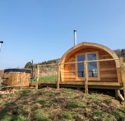 A wooden pod with a deck and railing next to a hot tub on a grassy hill at Holbein in Matlock