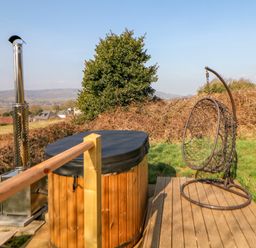 A wooden hot tub with a black cover and a metal chimney next to a hanging wicker chair on a wooden deck outside at Holbein in Matlock
