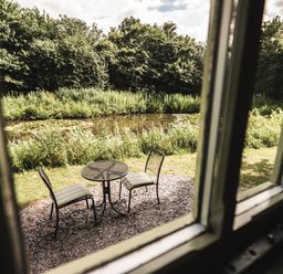 A small outdoor seating area with two striped chairs and a round table by a pond with greenery at Little Otchan Shepherd's Hut in Halsham near Roos