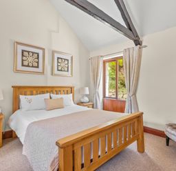 A bedroom with a wooden bed frame side tables lamps wall art a window with curtains and a chair at The Stables in St Neot