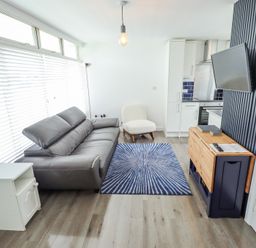 A living room with a gray sofa a white armchair a blue patterned rug a wooden cabinet a wall-mounted TV and kitchen area at 104 The Laurels in Chapel St Leonards