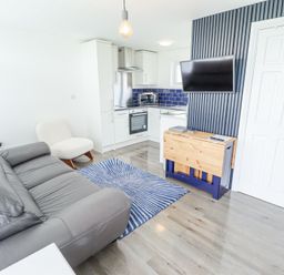 A living area with grey sofa white chair blue and white rug wall mounted tv and small kitchen at 104 The Laurels in Chapel St Leonards