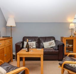 A seating area with a brown leather sofa and chairs a wooden table with a bottle and glasses and wooden cabinets with lamps at 5 Lakeview Rise Highampton near Hatherleigh