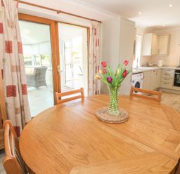 A dining area with a wooden table and a vase of flowers next to a kitchen with white cabinets and appliances at Bwthyn Clyd in Llanddaniel Fab