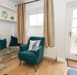 A corner of a room with a teal armchair with a cushion a stone seating area with cushions shelves with glass vases beige curtains a window and a glass door at Honeypot Cottage in Swanage
