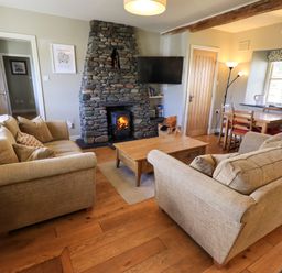 A living room with two beige sofas, a wooden coffee table, a stone fireplace, a wall-mounted TV, and a dining table with chairs at Fox Howe in Thornthwaite