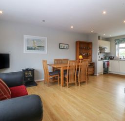 An open living area with a dining table and six chairs a small stove and a television next to a kitchen with white cabinets at Ty Nant in Abersoch