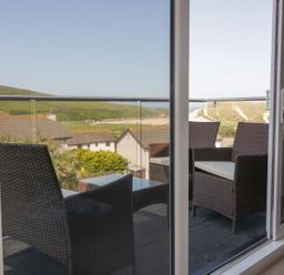 A balcony with wicker chairs and a glass table overlooking rooftops and hills at Atlantic View in Holywell Bay