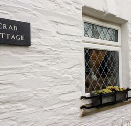 A white textured wall with a window featuring diamond-shaped panes and a flower box below it at Crab Cottage in Delabole