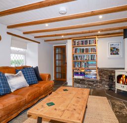 A living room with a leather sofa blue cushions wooden coffee table bookshelf and a wood burning stove at Crab Cottage in Delabole