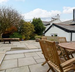 An outdoor patio with wooden chairs and table stone flooring and surrounding bushes at Crab Cottage in Delabole