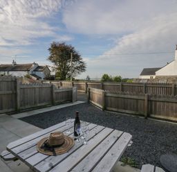 An outdoor wooden picnic table with a bottle and four glasses on a paved and gravel patio with a wooden fence and houses in the background at Edgemere Retreat in Cartmel