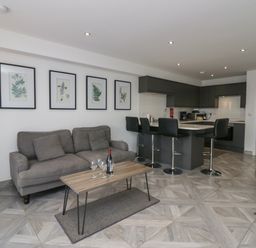A living room with a gray sofa a wooden coffee table with wine glasses and a bottle stools at a kitchen island with dark cabinets in Edgemere Retreat Cartmel