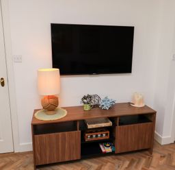 A wall-mounted television above a wooden cabinet with a lamp plant decor candles and books in a room at Coastal Retreat in Seahouses