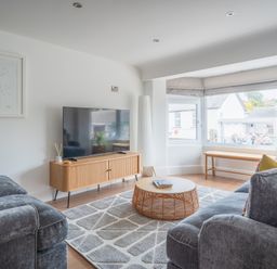 A living room with two grey sofas a wooden TV stand with a flat screen TV a round rattan coffee table a large window with bench seating and a colorful giraffe painting on the wall at Hoheck in Abersoch