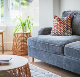 A grey sofa with a colorful pillow next to a woven side table with a green plant in a bright room at Hoheck in Abersoch