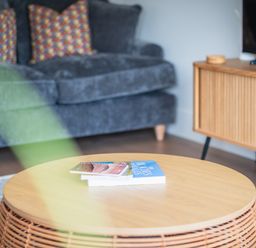 A round wooden coffee table with books in front of a grey sofa with patterned cushions and a wooden cabinet with a television at Hoheck in Abersoch