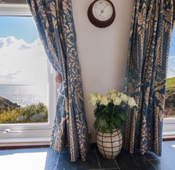 A corner with a vase of white flowers on the floor between two patterned curtains with a wall-mounted clock above with a view of hills and the sea through windows at Suntrap House in Portloe
