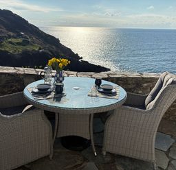 An outdoor patio with a round glass table and two wicker chairs overlooking the sea at Suntrap House in Portloe