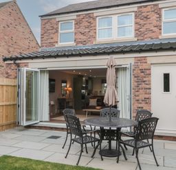 A patio with a round metal table and four chairs with an umbrella in front of a brick house with open folding doors at Breamish Christon Bank near Embleton