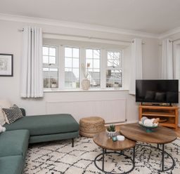 A living room with a green sectional sofa, wooden coffee tables, a television on a wooden stand, and windows with white curtains at The Palms in Abersoch