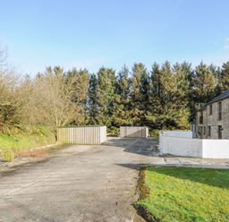 An outdoor area with a paved surface two wooden storage sheds a stone building and trees in the background at The Dairy Barn in Camelford