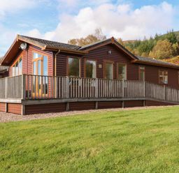 A wooden cabin with a fenced porch surrounded by grass and trees at Crathie Lodge in Banavie near Caol