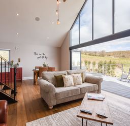 A living room with a beige sofa, coffee table, dining table, and large glass windows opening to a deck at Hare's Hill in Thornton-Le-Dale