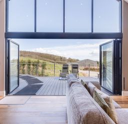 A living room with a sofa and pillows opening onto a deck with two chairs overlooking grass and a building at Hares Hill in Thornton-Le-Dale