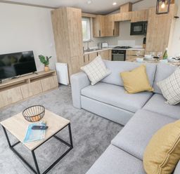 A living room with a grey sectional sofa and a wooden coffee table near a kitchen with wooden cabinets at Caldey Retreat in Hasguard Cross near Broad Haven