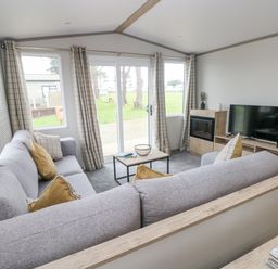 A living room with grey sofas, a coffee table, a TV on a wooden stand, and large windows with checkered curtains at Caldey Retreat Hasguard Cross near Broad Haven