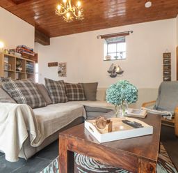 A living room with a gray sectional sofa and pillows a wooden coffee table with a tray and flowers a gray chair with a blanket a wooden ceiling and white walls with a small window at Owl Cottage in Ashen Cross Pelynt