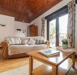 A living room with a sofa, cushions, wooden coffee table with a plant, wooden floor, and glass doors with floral curtains at Court Cottage in Ashen Cross, Pelynt