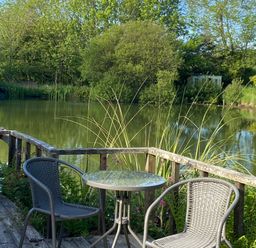An outdoor seating area with a small glass table and two chairs on a wooden deck by a pond with trees in the background at Court Cottage in Ashen Cross Pelynt