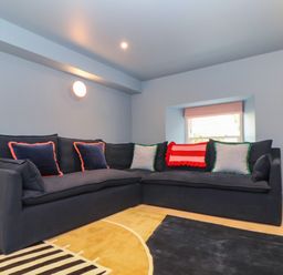 A corner sofa with cushions in a living room with a lamp and a window at Mavericks Wave in St Agnes