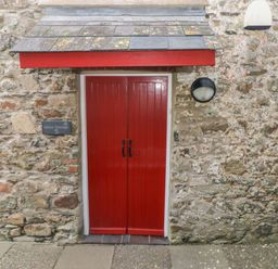 A red double door entrance with black handles under a small roof on a stone wall at Wood Cottage in Solva