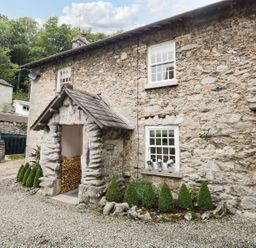 A stone cottage exterior with a wood storage area and small shrubs at Pear Tree Cottage in Grange-Over-Sands